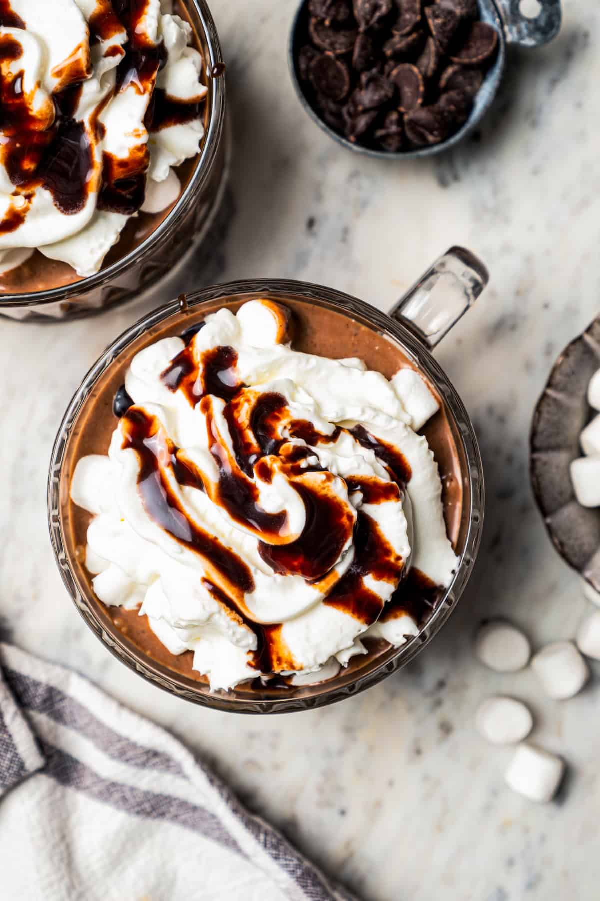 Overhead image of two glass mugs with hot chocolate topped with whipped cream and a drizzle of chocolate sauce.