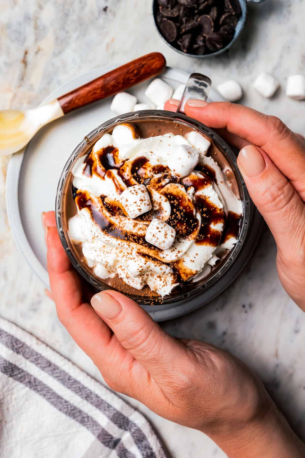 Two hands wrapped around a mug of dark hot chocolate topped with whipped cream and chocolate sauce, next to a small spoon and scattered mini marshmallows.