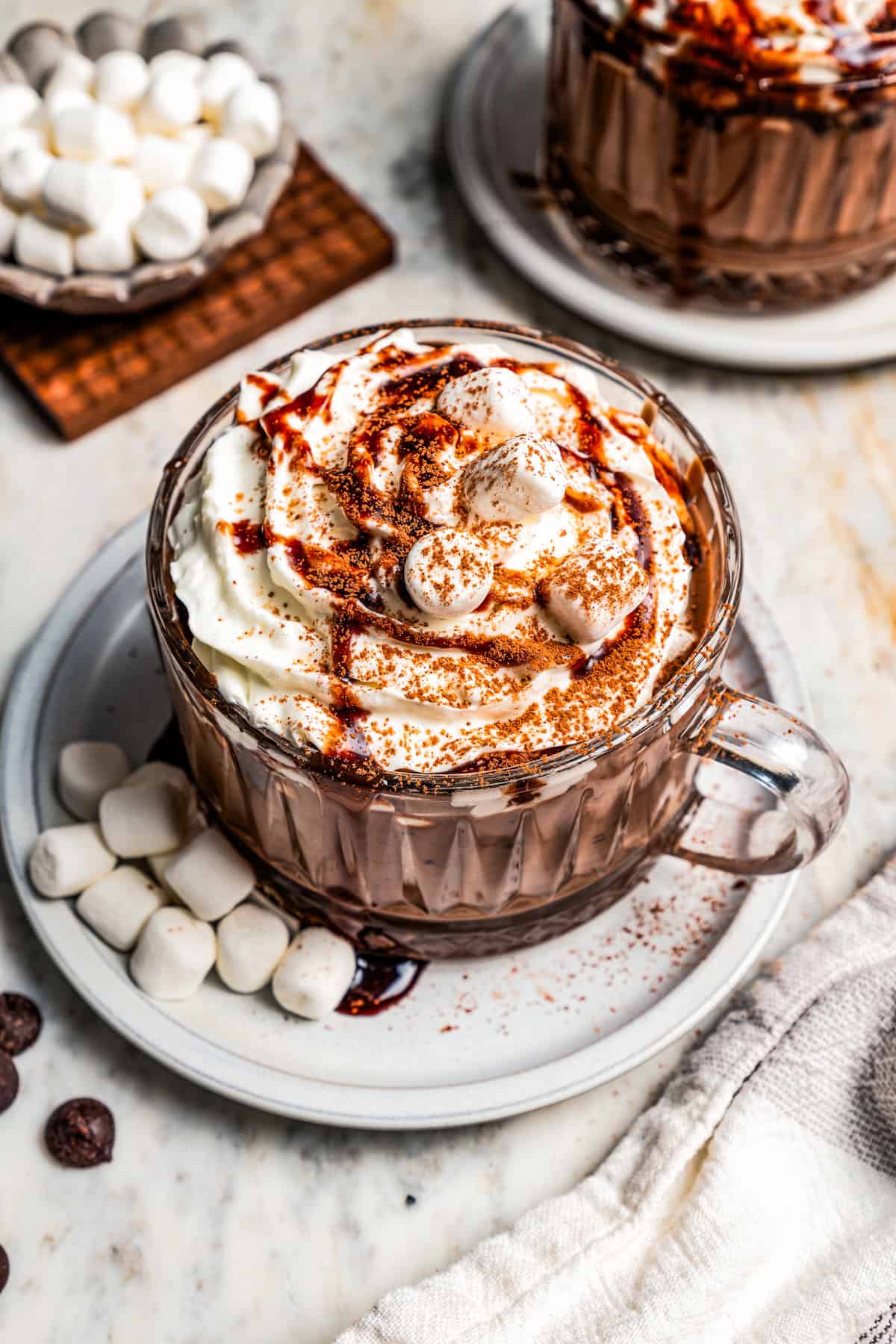 Dark hot chocolate in a glass mug topped with whipped cream and chocolate sauce, resting on a saucer next to mini marshmallows, with more marshmallows and a second mug in the background.