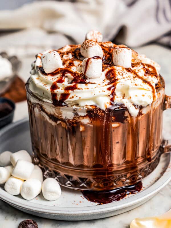 Dark hot chocolate in a glass mug topped with whipped cream and chocolate sauce, resting on a saucer next to mini marshmallows.