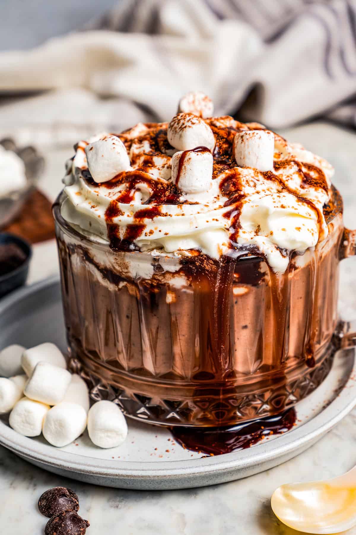 Dark hot chocolate in a glass mug topped with whipped cream and chocolate sauce, resting on a saucer next to mini marshmallows.