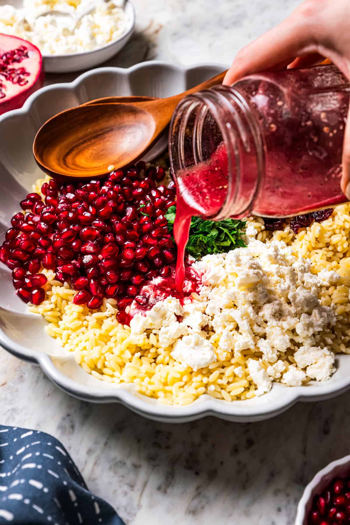 Pouring vinaigrette dressing from a mason jar over orzo pasta salad ingredients in a bowl.