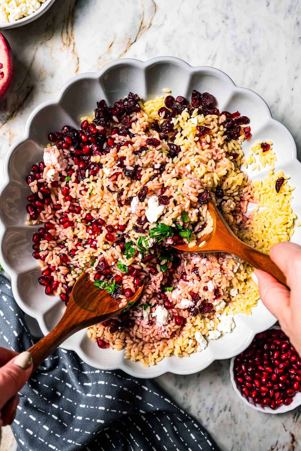 Using two spoons to combine orzo, pomegranate arils, cheese, and herbs in a bowl.