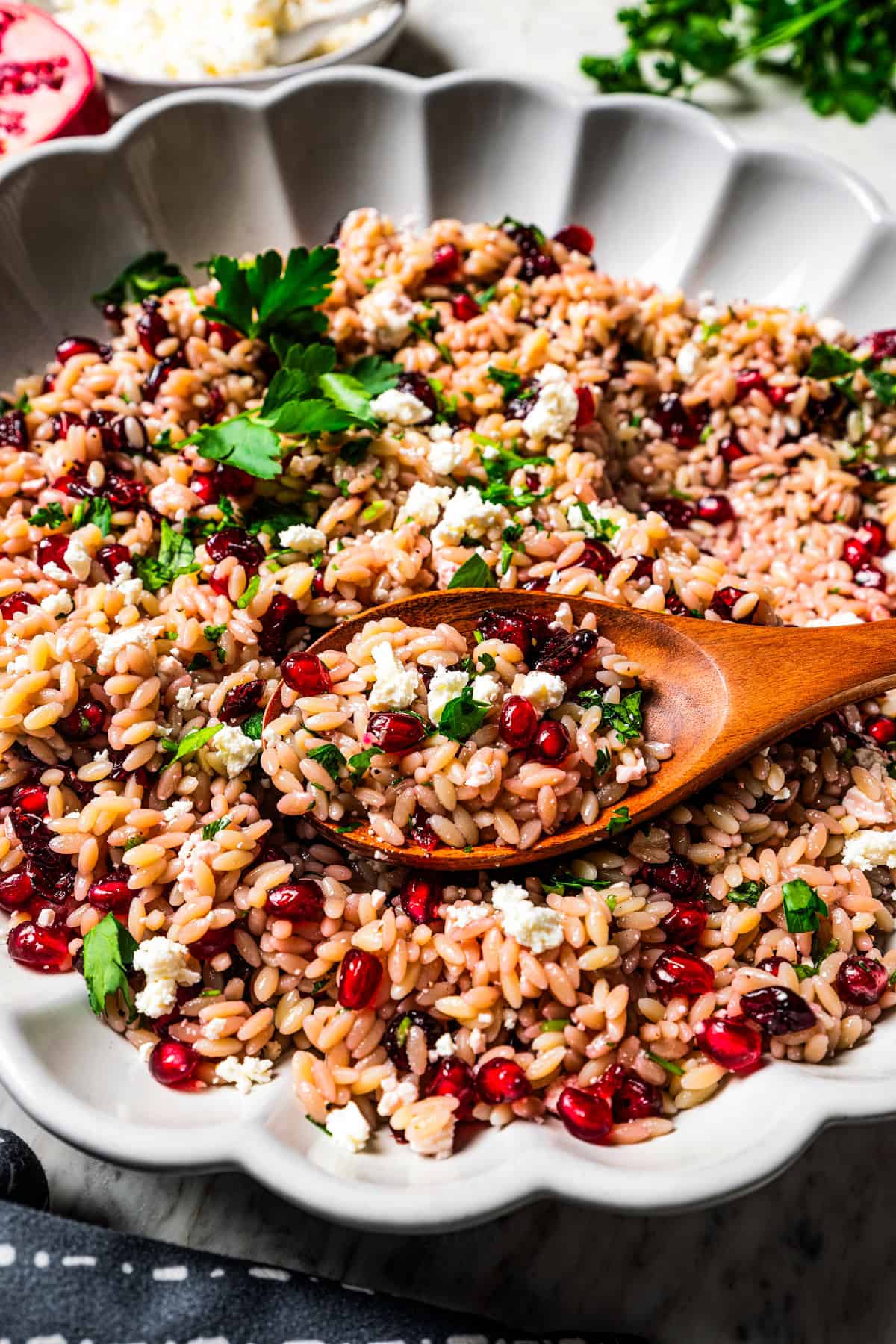 Wooden spoon stirring through orzo salad with pomegranate arils and cheese.