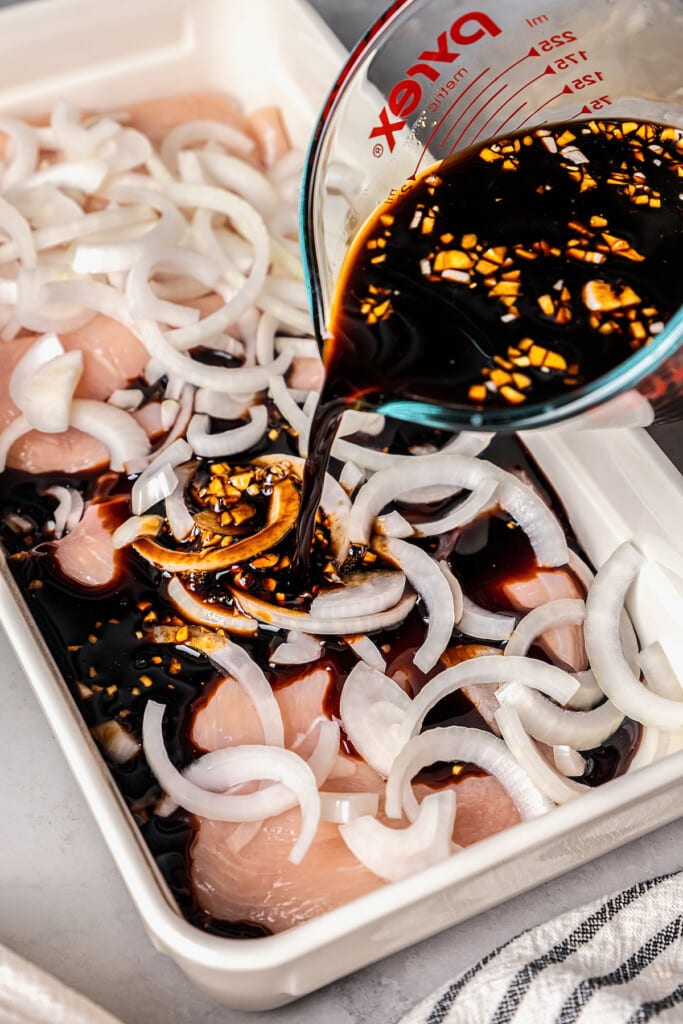 Teriyaki marinade poured over uncooked chicken breasts topped with onion slices in a baking dish.