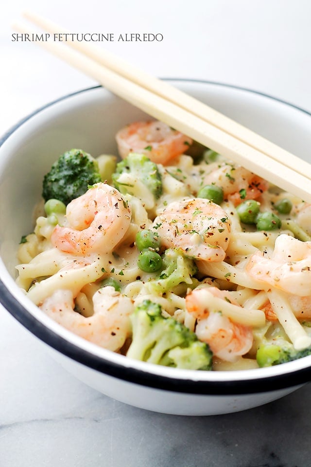 A white bowl with blue rim, filled with fettucine pasta, shrimp, broccoli, and peas in a creamy sauce.
