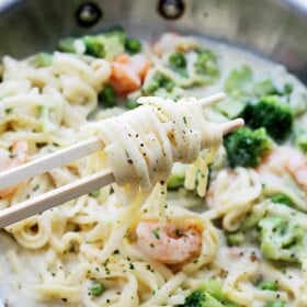 Pasta alfredo with shrimp and vegetables in a skillet. A pair of chopsticks is swirling a bite-sized portion of pasta out of the skillet.