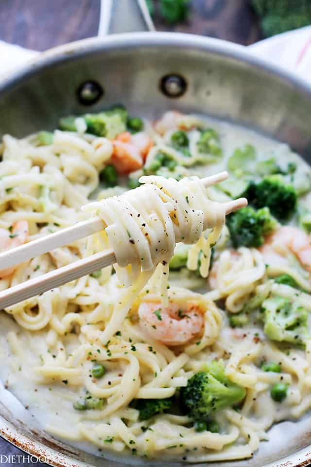 Pasta alfredo with shrimp and vegetables in a skillet. A pair of chopsticks is swirling a bite-sized portion of pasta out of the skillet.