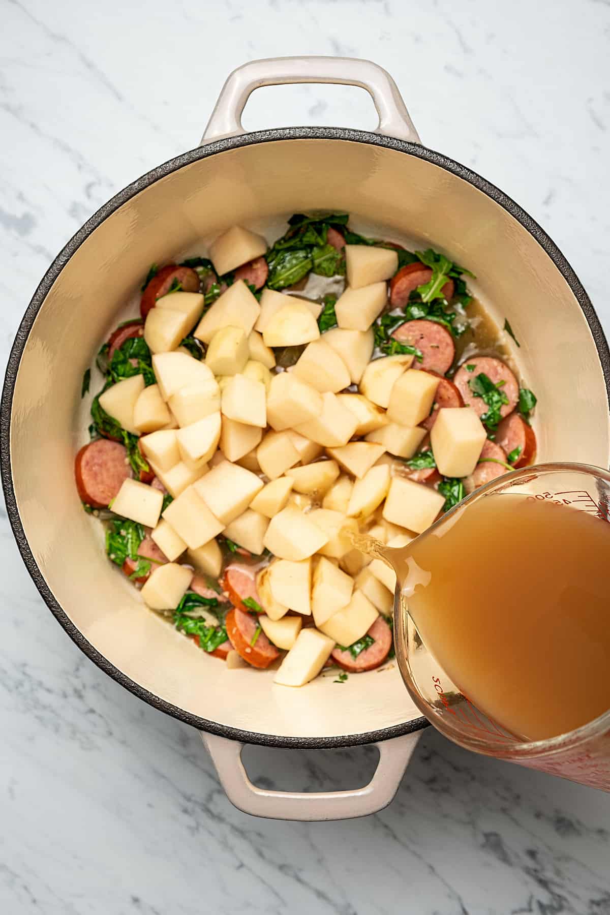 Pouring chicken broth over potatoes, kale, and sausage in a Dutch oven.