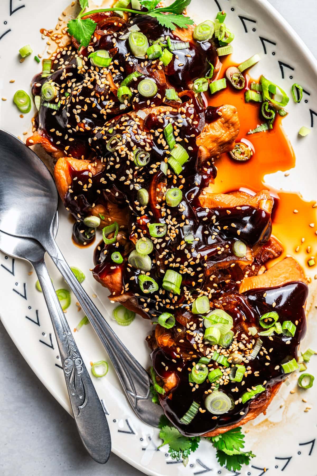 Overhead image of baked teriyaki chicken served on a white platter and covered in sauce, next to serving spoons.