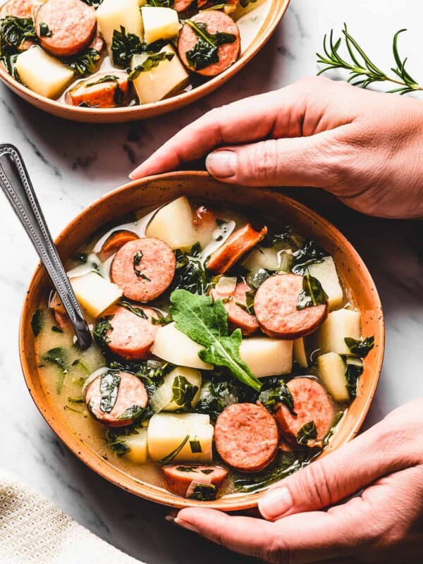 hands picking up a bowl of potato soup with kale and sausages.
