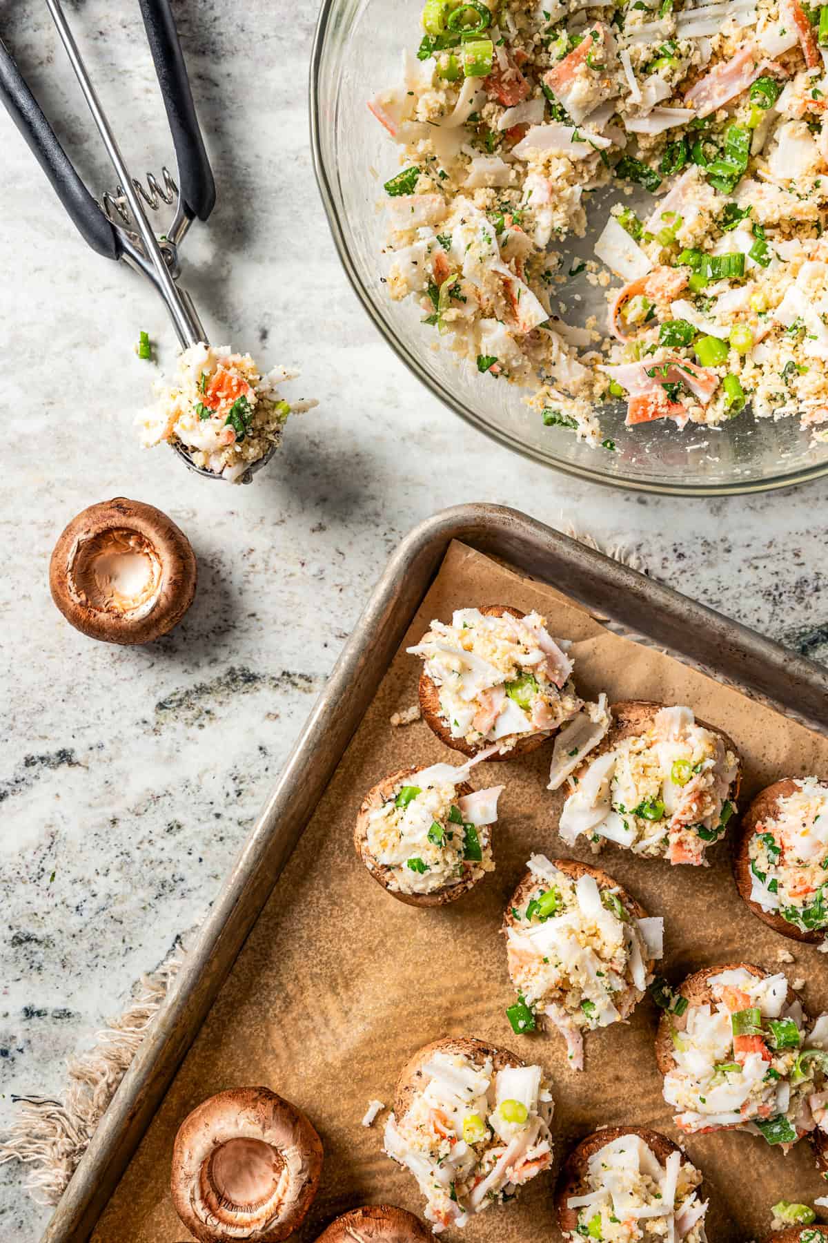 Partially stuffed mushrooms on the corner of a lined baking sheet, next to a small cookie scoop and a bowl of crab filling.