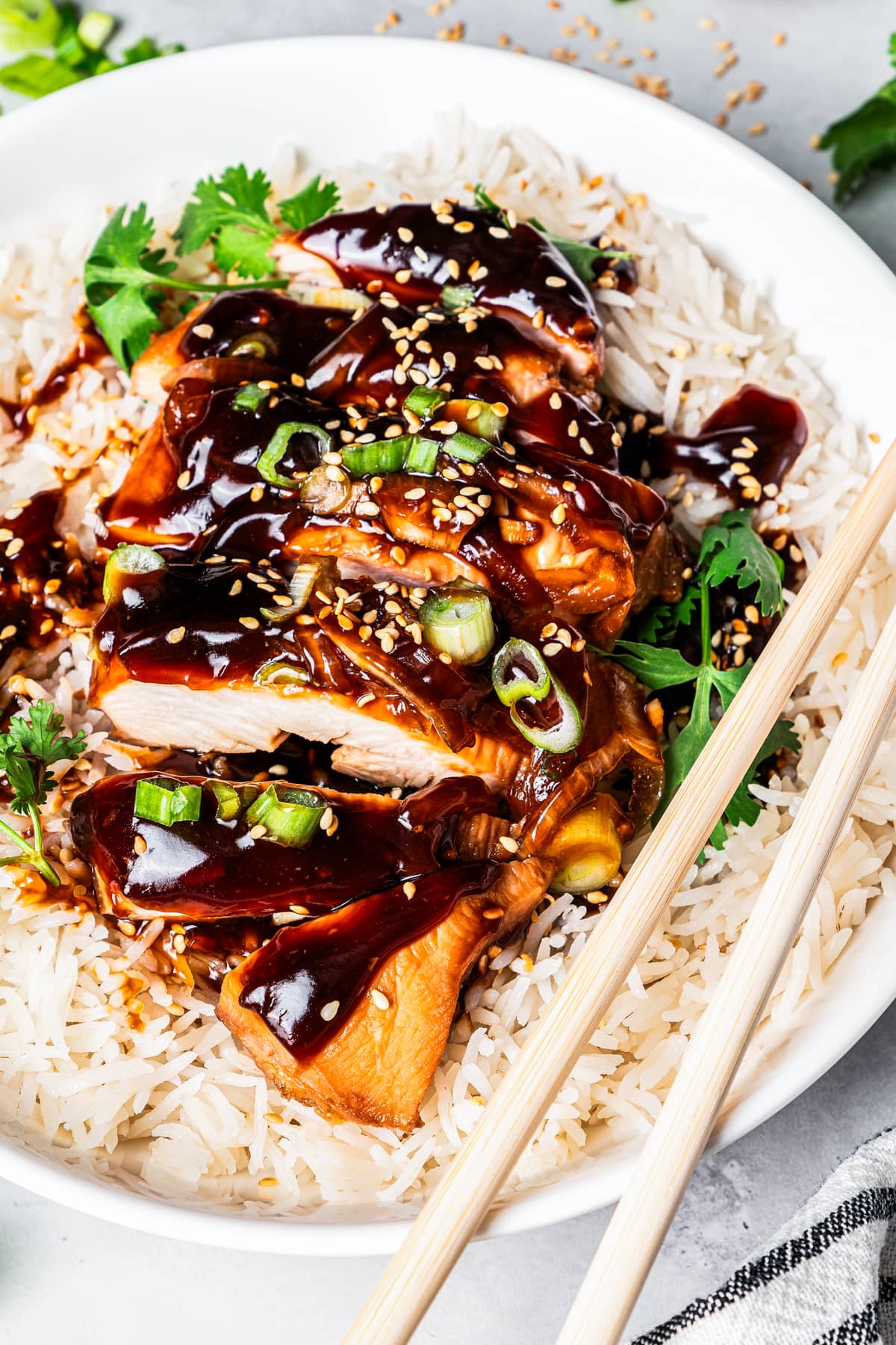 Close-up overhead view of chicken teriyaki, sliced, and served over rice on a white plate, garnished with green onions and sesame seeds, with a set of chopsticks.