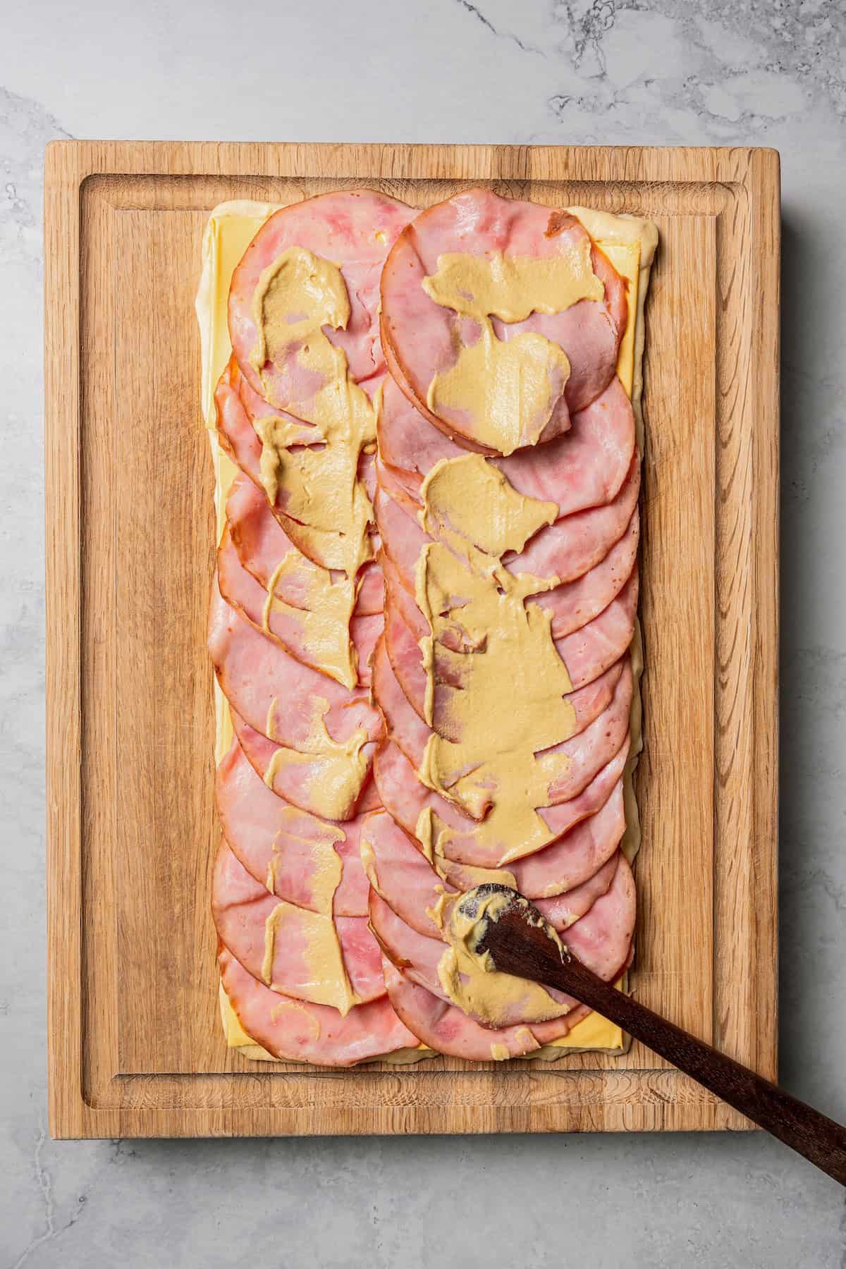 A knife spreading mustard over ham and cheese layered over crescent roll dough on a wooden cutting board.