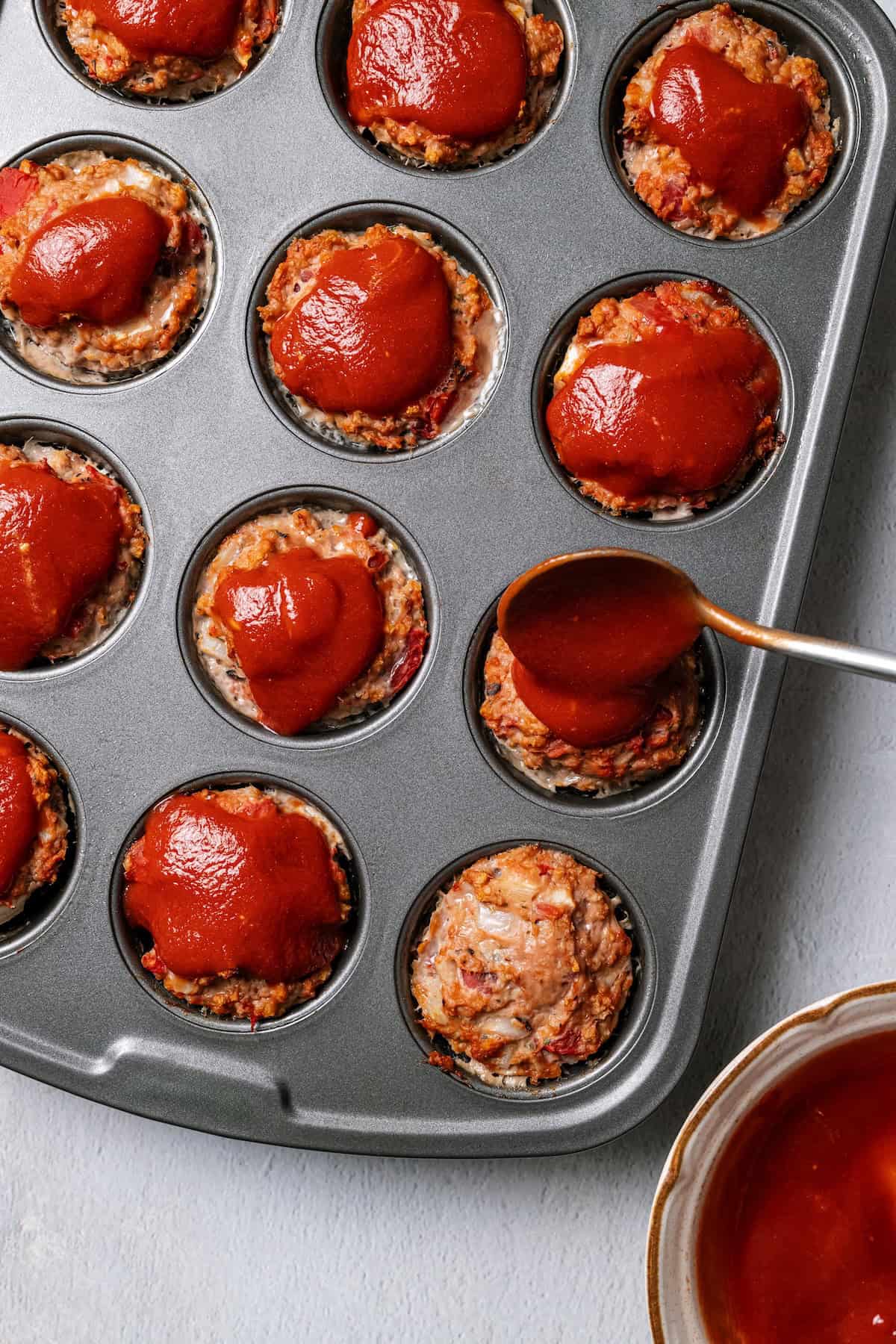 Overhead view of a spoon adding sauce over mini turkey meatloaves in a metal muffin tin.