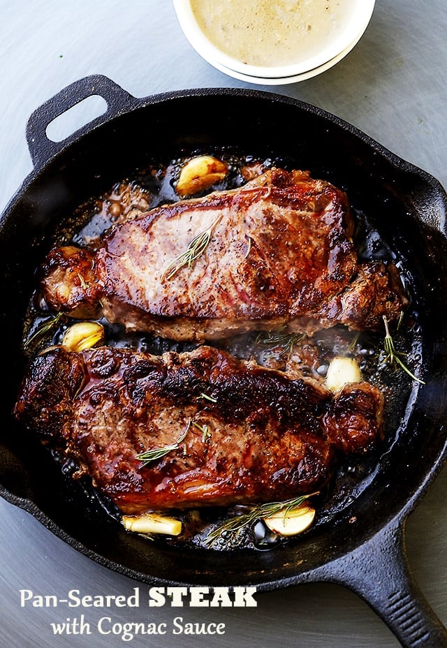 Overhead view of two pan seared steaks in a cast iron skillet with garlic cloves and rosemary, next to a bowl of cognac sauce.