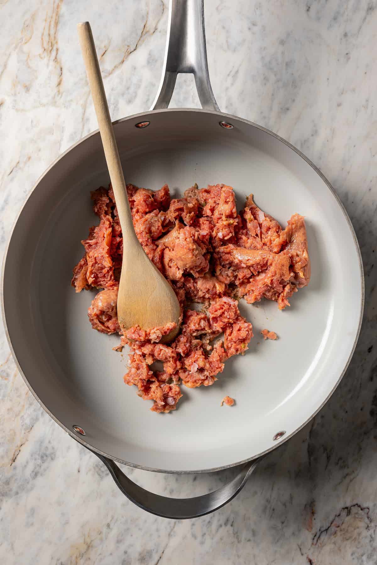 Ground Italian sausage broken up in a skillet with a wooden spoon.