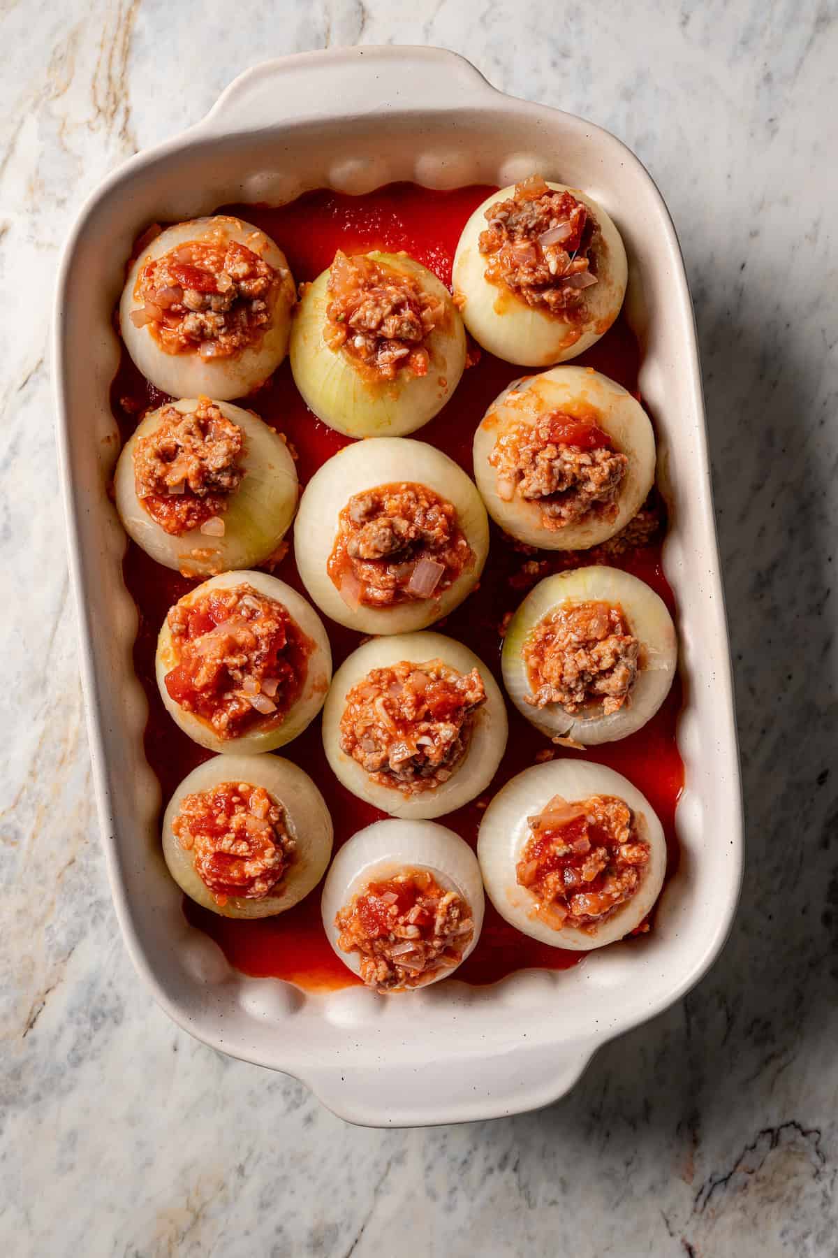 Sausage stuffed onions arranged over a layer of tomato sauce inside a baking dish.