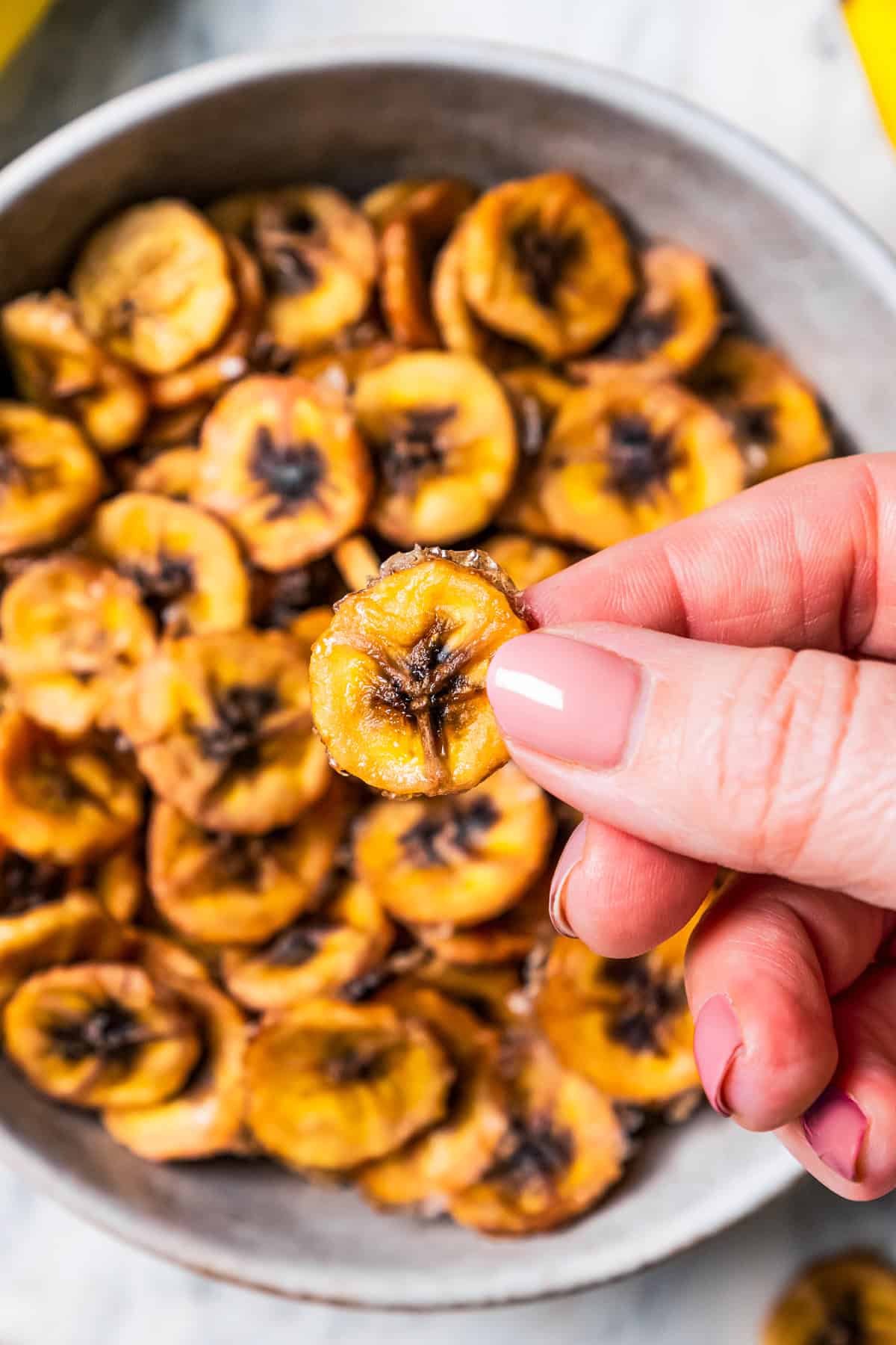 A hand holding up a baked banana chip over more banana chips in a bowl.