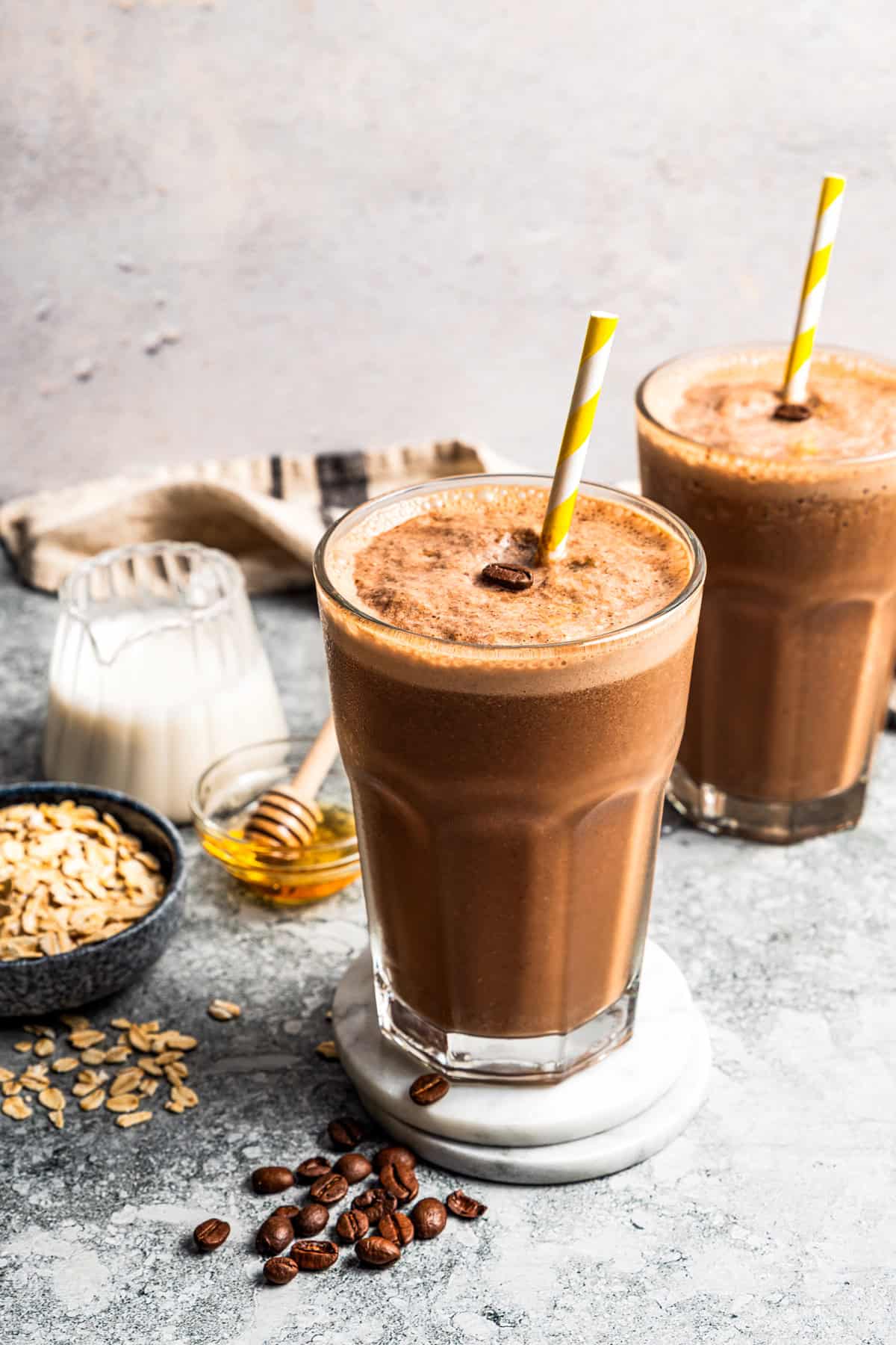 A coffee smoothie in a tall glass with a straw and a second smoothie in the background.