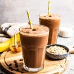 A coffee smoothie in a tall glass with a straw and a second smoothie next to a bowl of oats in the background.