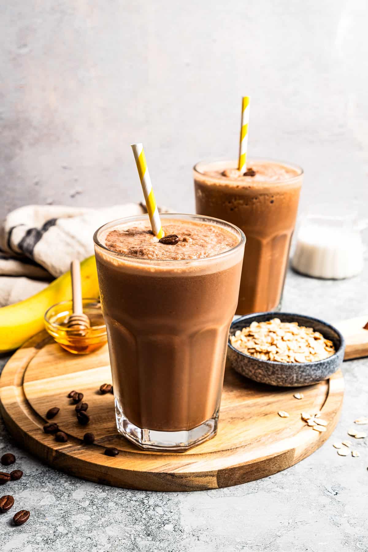 A coffee smoothie in a tall glass with a straw and a second smoothie next to a bowl of oats in the background.