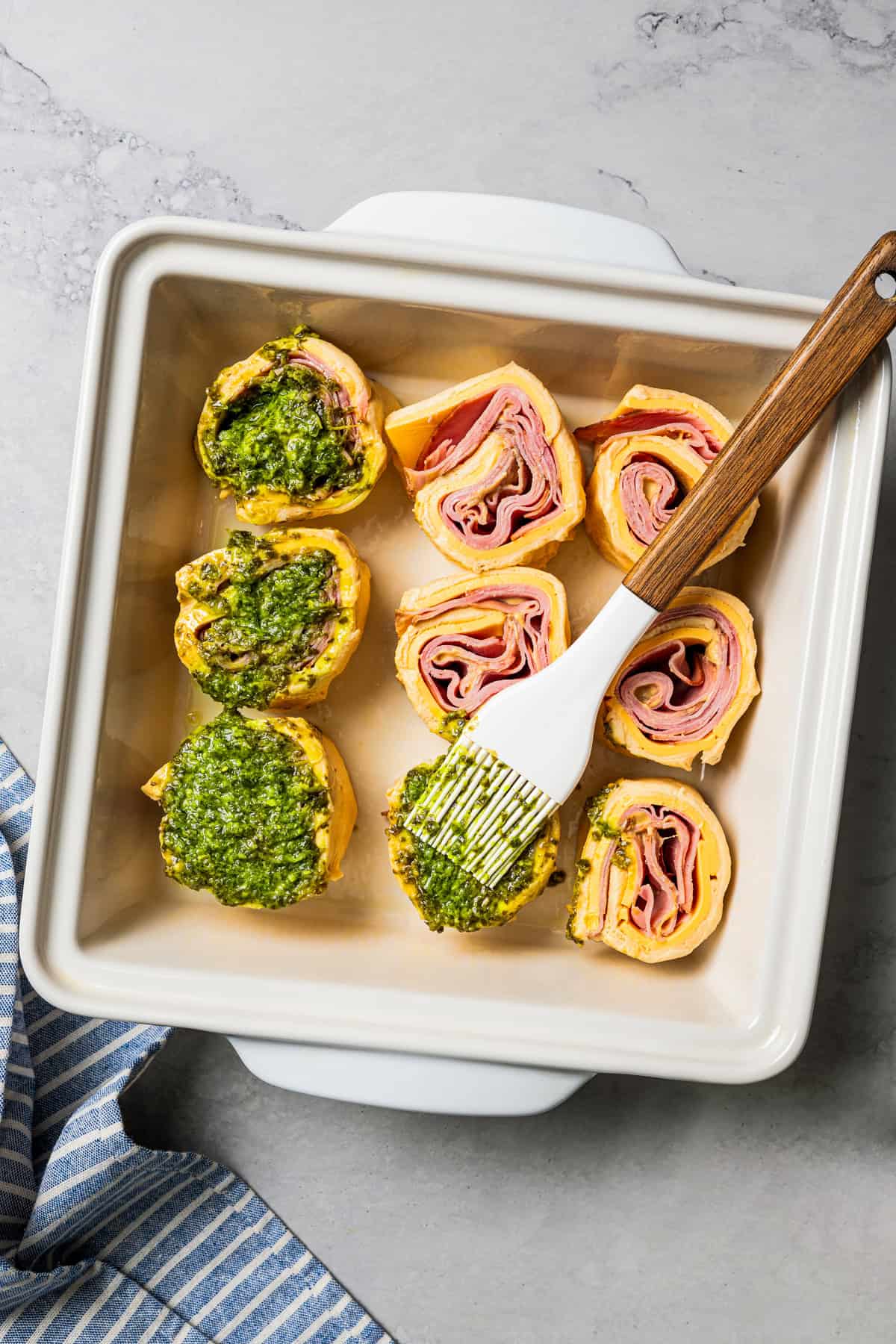 A basting brush coating the tops of roll ups with basil pesto in a baking dish.