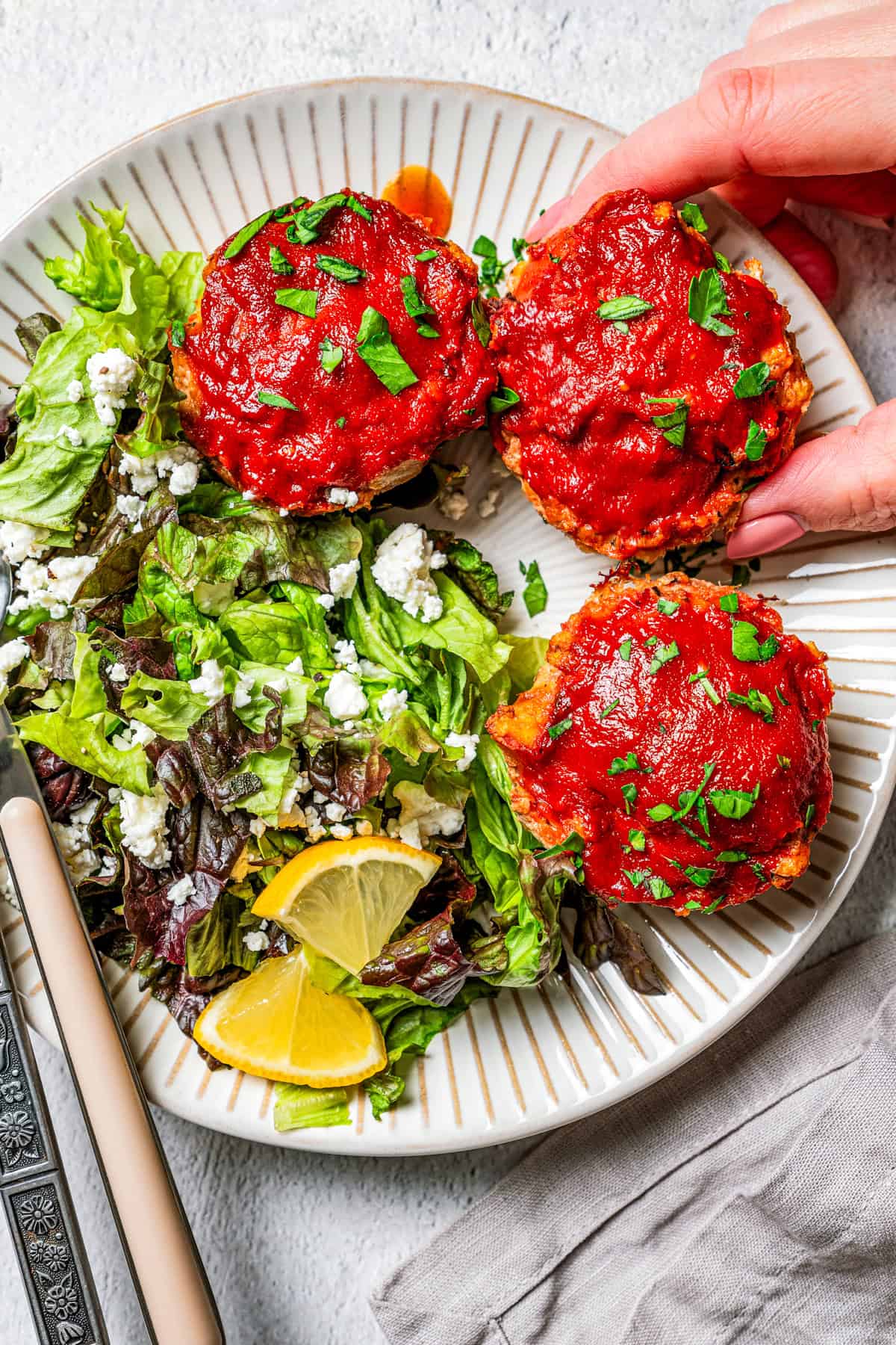 A hand picking up one of three mini turkey meatloaves next to a side of salad on a white plate.