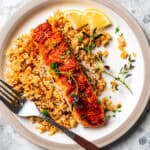 Salmon filet served over a bed of rice on a plate next to a fork and lemon wedges.