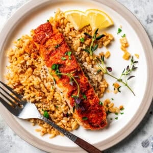 Salmon filet served over a bed of rice on a plate next to a fork and lemon wedges.