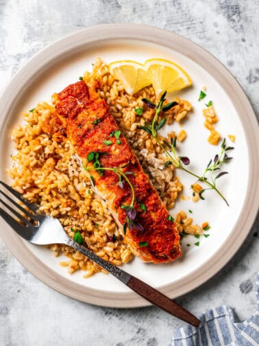 Salmon filet served over a bed of rice on a plate next to a fork and lemon wedges.