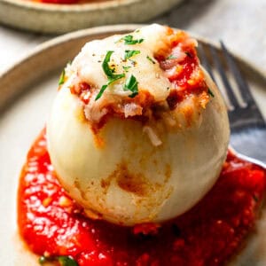 A stuffed onion served over tomato sauce on a dinner plate next to another plate.