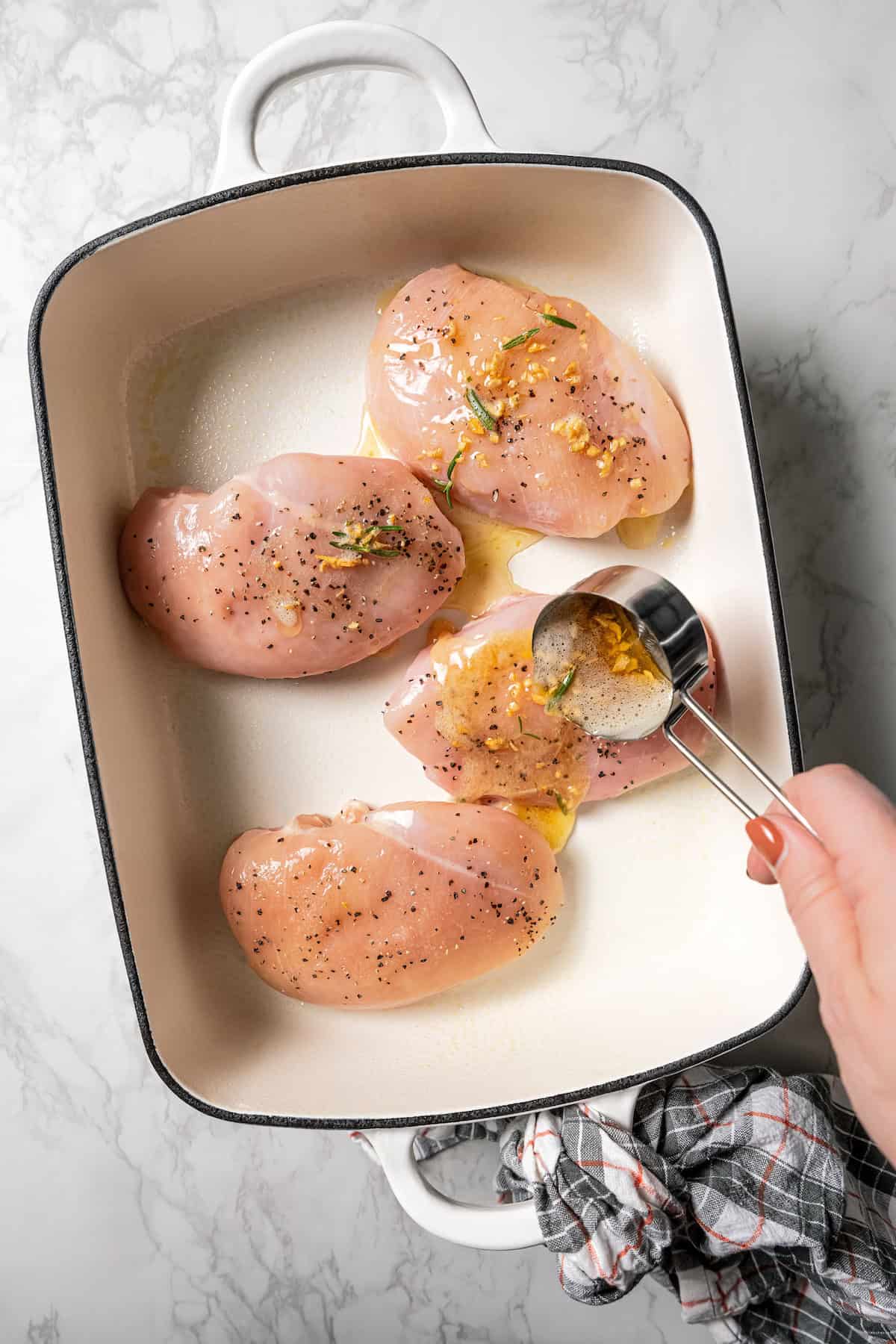 A hand spooning garlic butter sauce over chicken breasts in a ceramic baking dish.