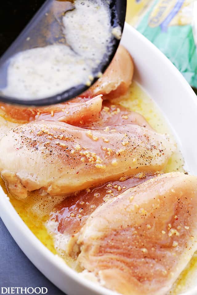 Pouring garlic butter sauce over four raw chicken breasts in a white baking dish.