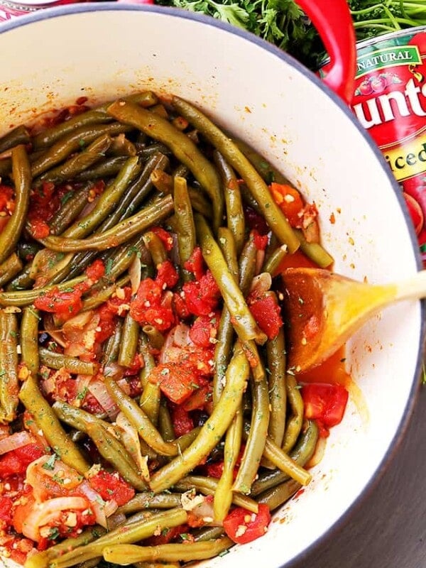 Overhead view of braised green beans and tomatoes in a pot with a wooden spoon.