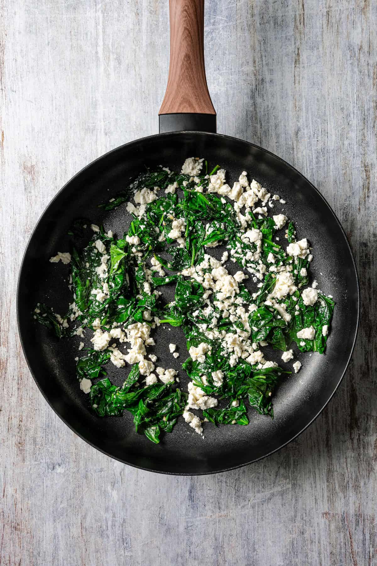 Feta cheese added to sautéed spinach in a skillet.