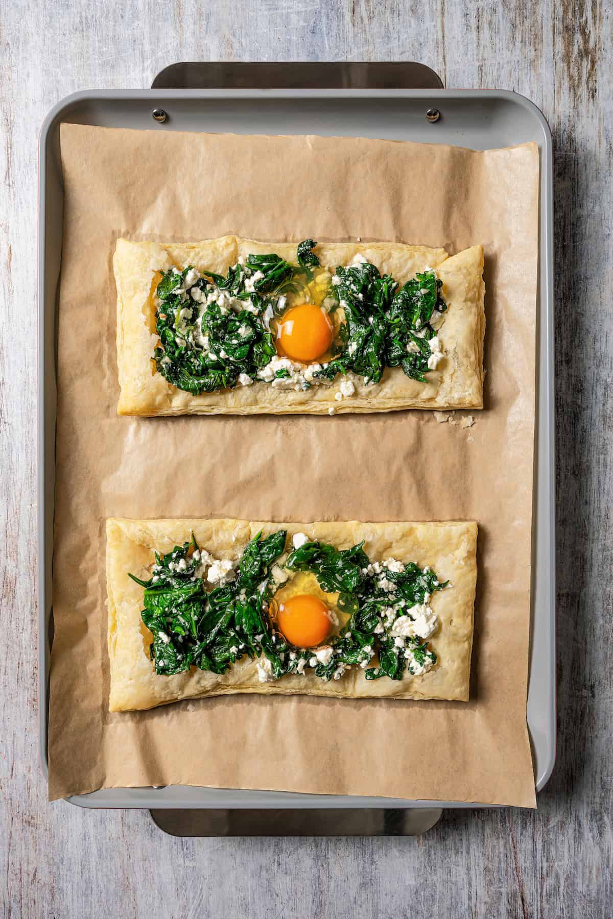 Two puff pastry sheets topped with spinach and feta mixture and an egg on a parchment-lined baking sheet.