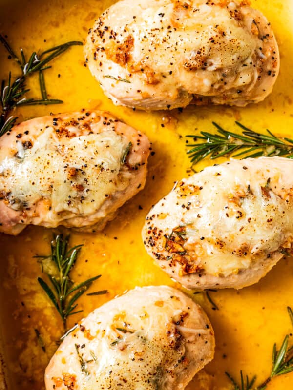 Up close shot of garlic butter chicken topped with melted cheese next to rosemary sprigs in a baking dish.