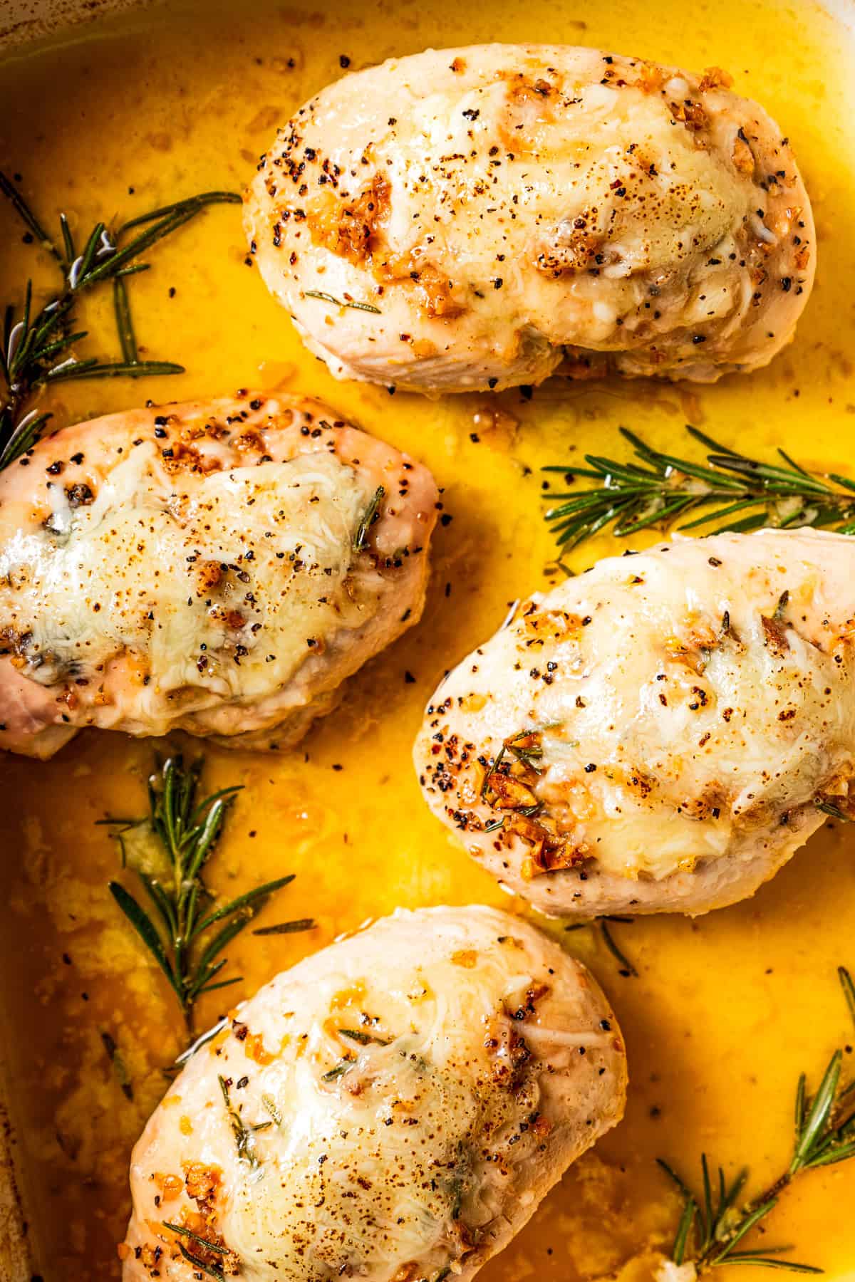 Up close shot of garlic butter chicken topped with melted cheese next to rosemary sprigs in a baking dish.