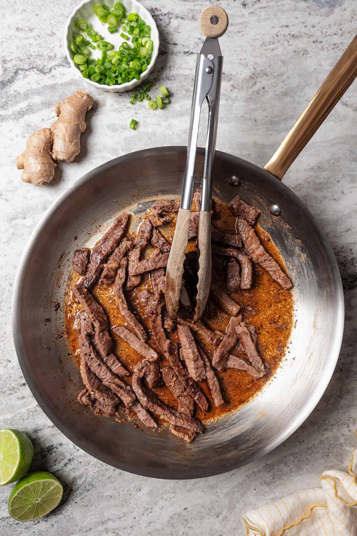 Beef strips combined with ginger stir fry sauce in a metal skillet with a set of tongs.