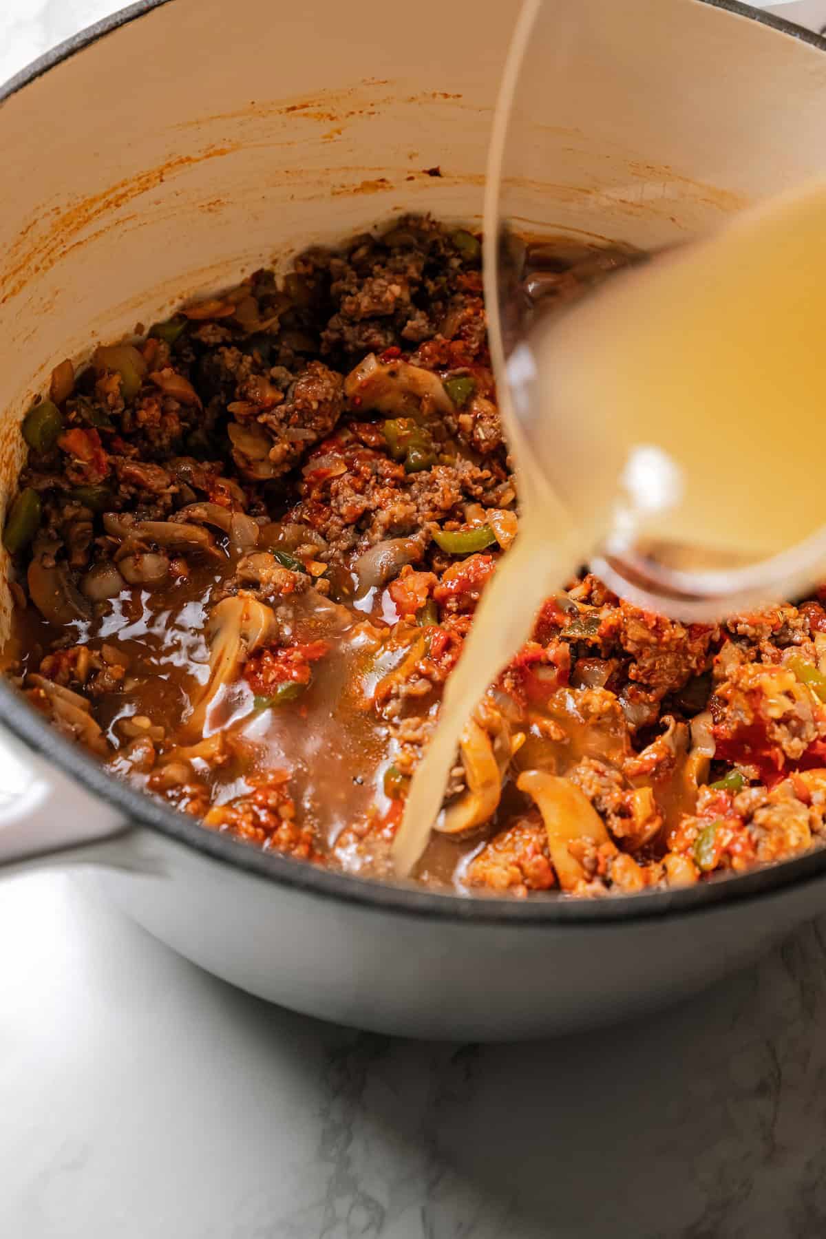 Chicken stock being poured into a large pot of sausage spaghetti sauce.