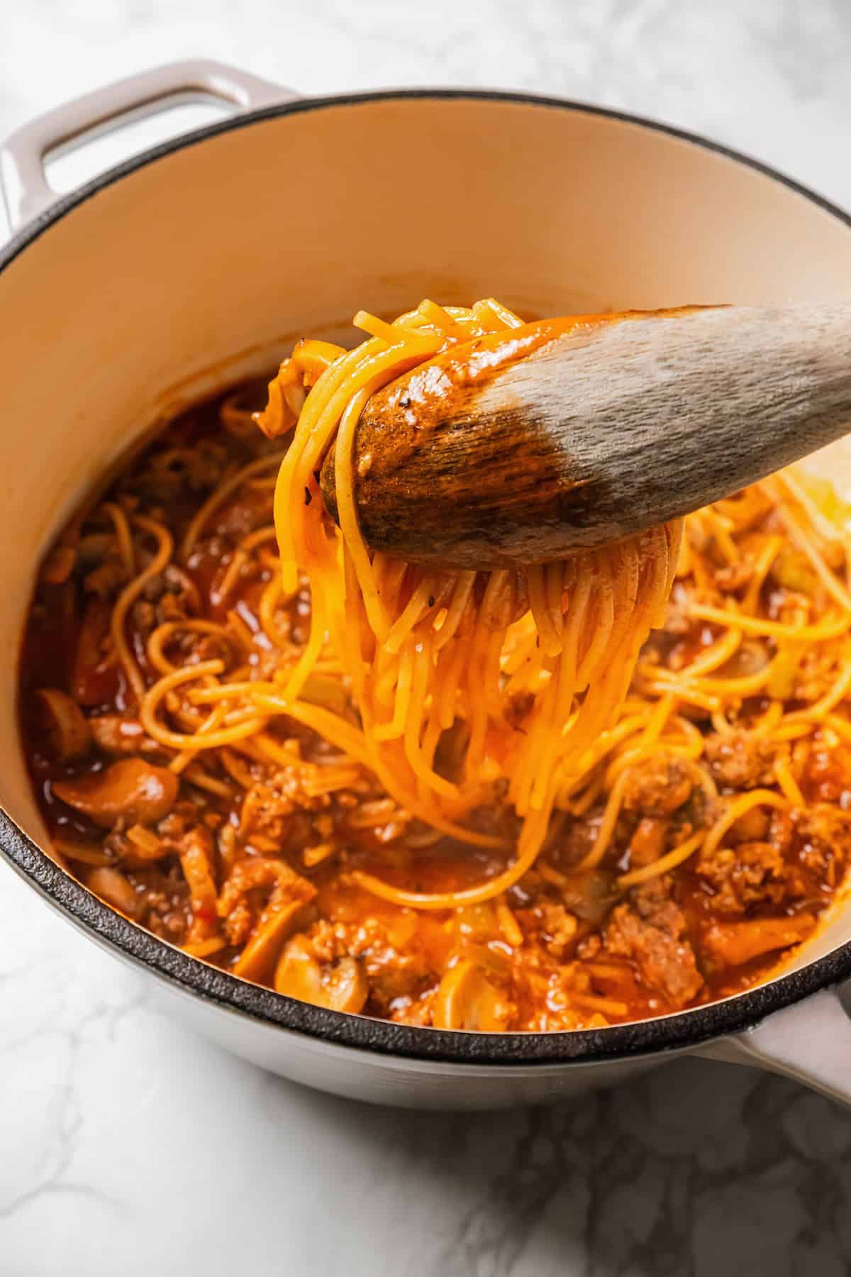 A set of wooden tongs lifting sausage spaghetti from a large pot.