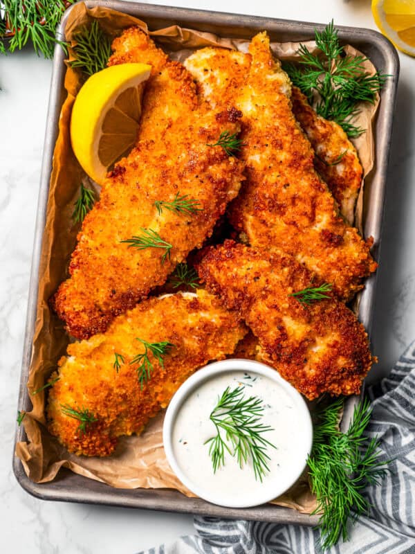 Overhead view of parmesan chicken tenders garnished with lemon wedges on a tray next to a small bowl of buttermilk ranch dressing.