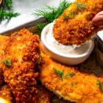 Dipping a parmesan chicken tender into a small bowl of buttermilk ranch dressing next to more chicken tenders on a tray.