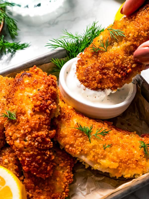 Dipping a parmesan chicken tender into a small bowl of buttermilk ranch dressing next to more chicken tenders on a tray.