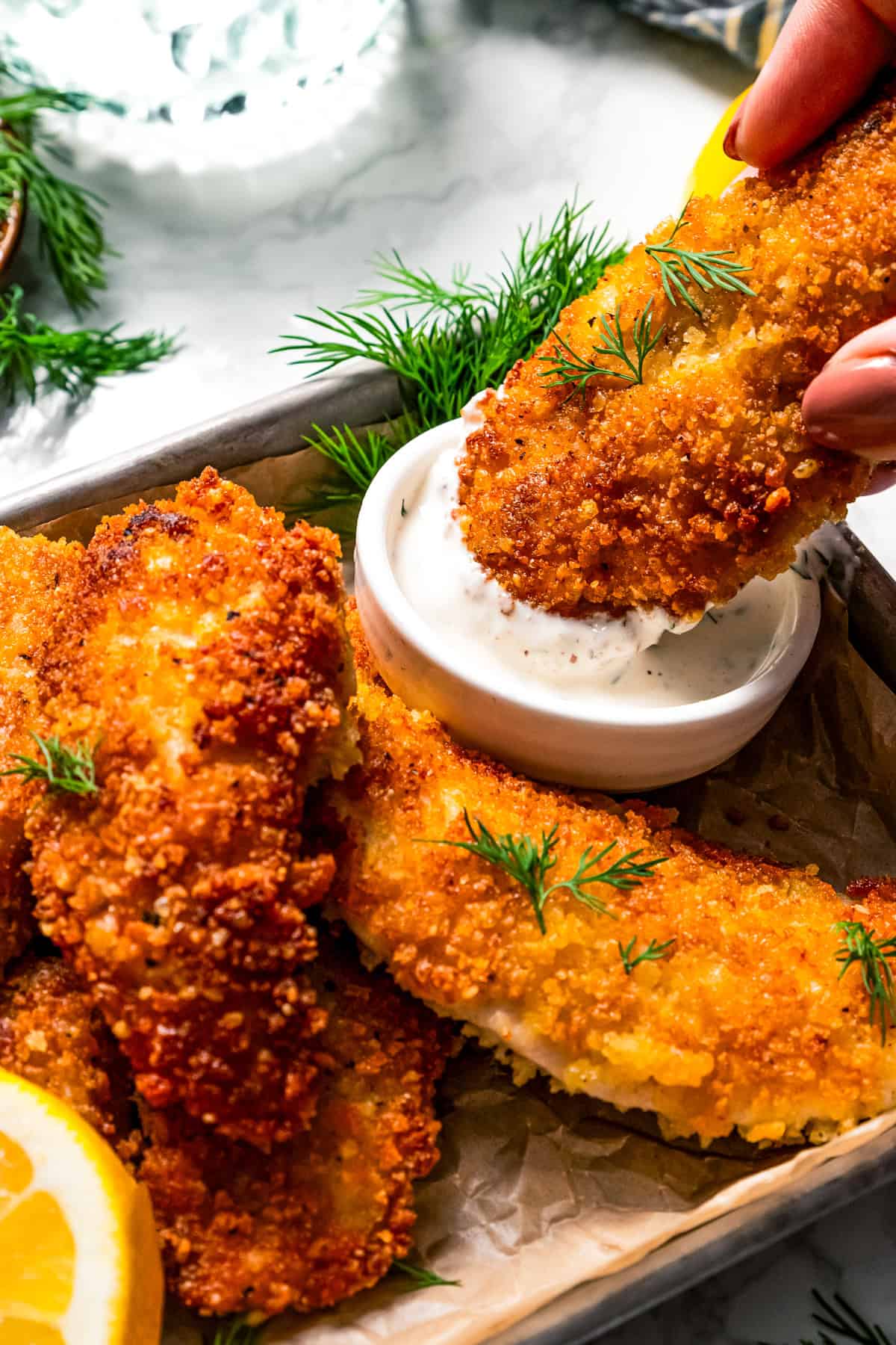 Dipping a parmesan chicken tender into a small bowl of buttermilk ranch dressing next to more chicken tenders on a tray.