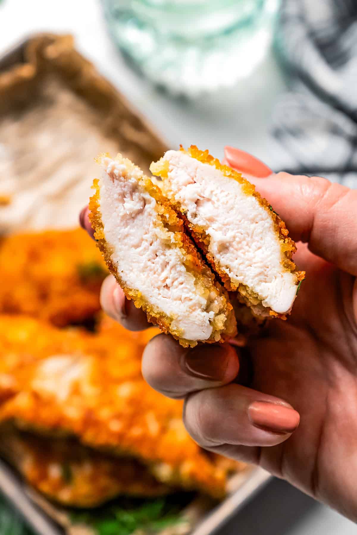 A hand holding up two halves of a parmesan chicken tender to expose the interior, with a tray of chicken tenders in the background.