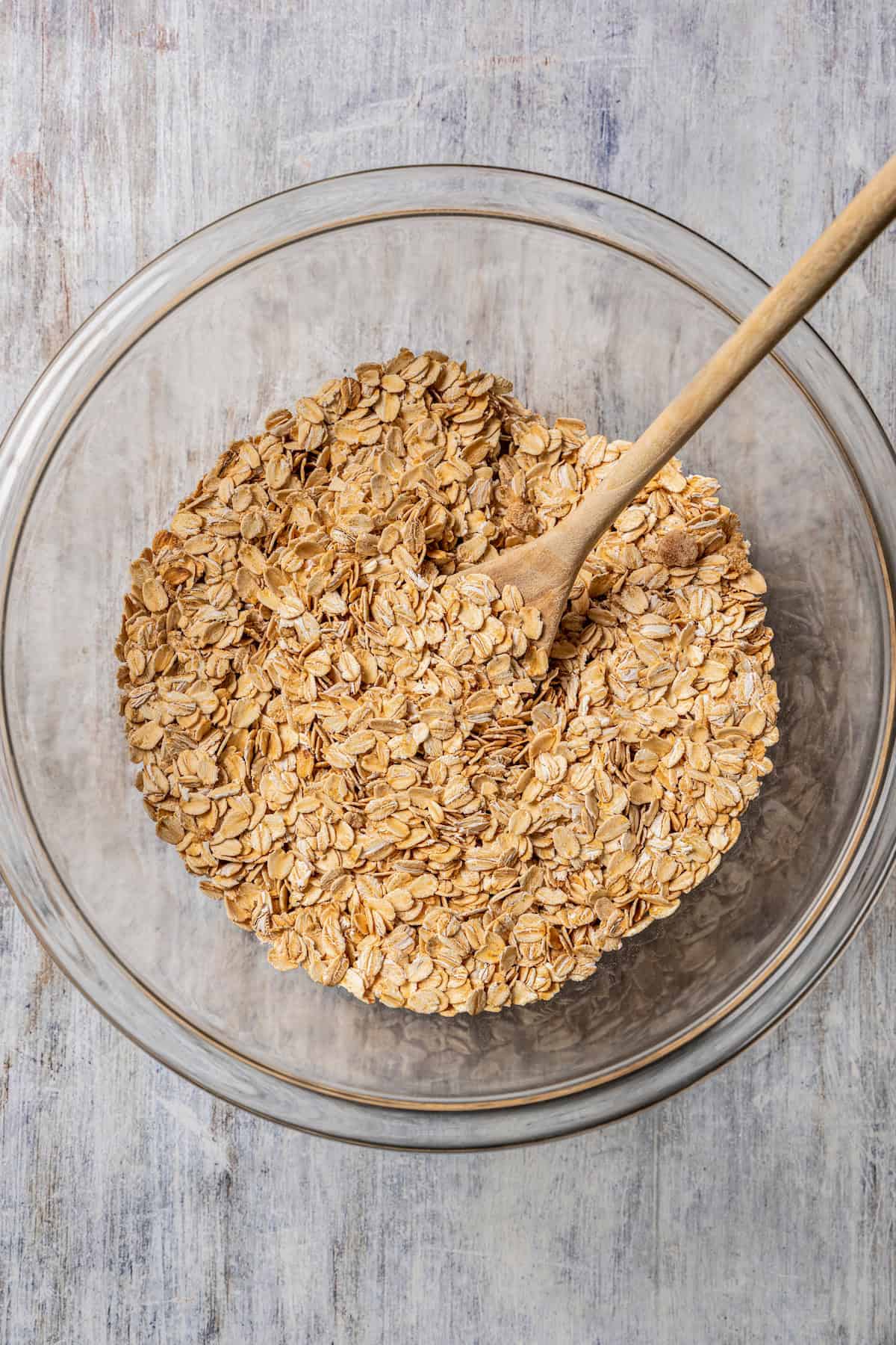 Oats and dry ingredients combined in a glass bowl with a wooden spoon.