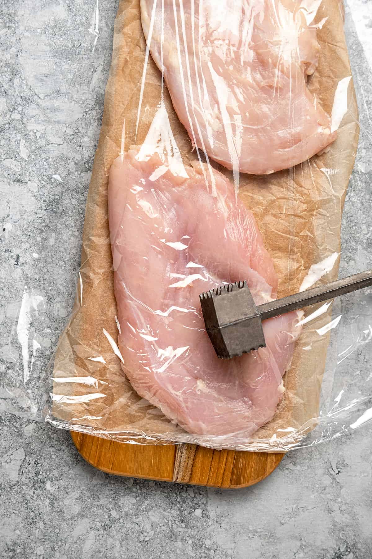 A mallet is used to flatten chicken breasts inside a sealed plastic bag on top of a wooden board.