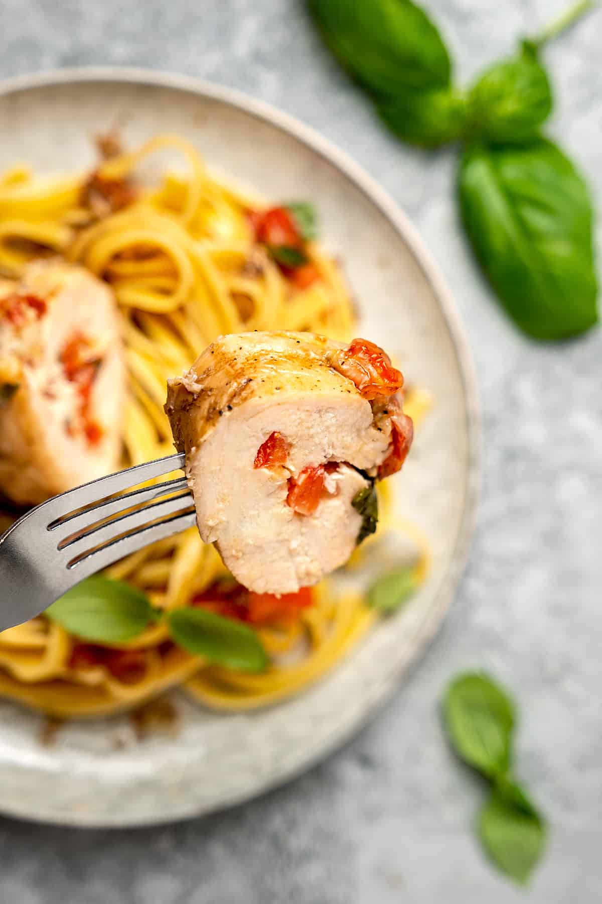 A fork holds up a slice of bruschetta stuffed chicken breast with a plate of pasta in the background.